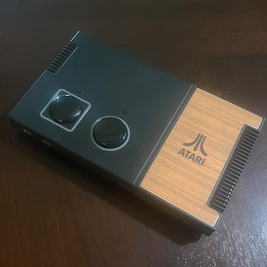 Puede incluir: Una consola de videojuegos Atari negra con un panel de madera en el lateral. La consola tiene dos botones redondos y el logotipo de Atari está impreso en el panel de madera.