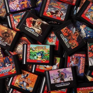 May include: A pile of Sega Genesis video game cartridges. The cartridges display vibrant artwork and game titles including Sonic 3, Streets of Rage, and Shining Force. The cartridges are black with red and white labels.