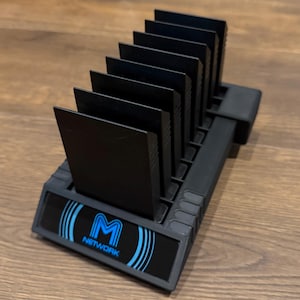 Könnte beinhalten: Ein schwarzer Kunststoff-Kassettenhalter mit Schlitzen, der mehrere schwarze Spielkassetten aufnimmt. Die Basis des Halters trägt das Wort "NETWORK" unter einem blauen "M"-Logo mit geschwungenen Linien.