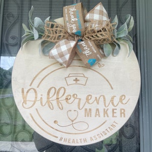 May include: A round wooden sign with the text "Difference Maker" and a stethoscope graphic. The sign is decorated with a burlap bow, a white and brown checked bow, and a blue ribbon with the text "Thank you" and "Heroes work here". The sign also includes the text "#Health Assistant".
