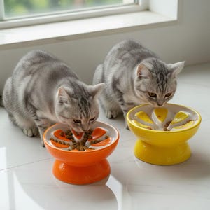 May include: Two gray tabby cats eating from ceramic slow feeder bowls. One bowl is orange, the other is yellow. The bowls have a raised design to slow down eating. The bowls are on a white tile floor.
