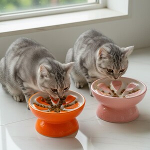 May include: Two gray tabby cats eating from ceramic slow feeder bowls. One bowl is orange, the other is pink. The bowls have a flower-like design with raised sections to slow down eating. The cats are on a white tiled floor.
