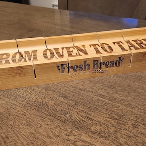 May include: A light brown bamboo bread slicer with the words "FROM OVEN TO TABLE" and "Fresh Bread" etched into the wood. The slicer has slots for guiding a knife and a curved, oval shape.