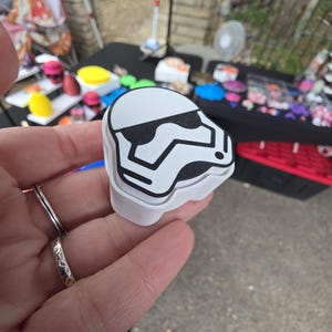 Può includere: Un oggetto bianco e nero a forma di casco da Stormtrooper. L'oggetto ha un design a strati con un contorno nero e dettagli neri sul casco. L'oggetto è tenuto in mano, con altri oggetti visibili sullo sfondo.