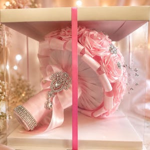 Op de afbeelding: Een boeket van lichtroze satijnen lintrozen in een doorzichtige vierkante displaydoos. Het handvat is omwikkeld met bijpassend roze satijnen lint, versierd met een zilverkleurige strass-broche en een band van fonkelende heldere kristallen.