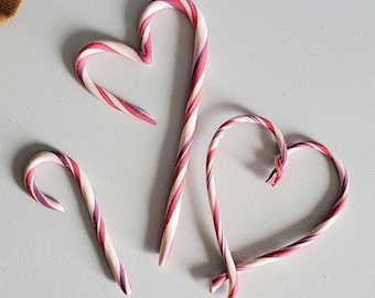 Puede incluir: Tres bastones de caramelo con forma de corazón. Los bastones de caramelo son rojos y blancos con un remolino rosa.