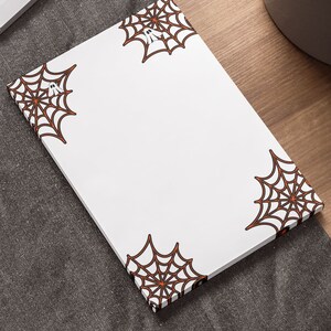 May include: White notepad with a black and orange spiderweb design on each corner. The notepad is on a gray fabric surface.