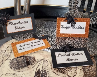 May include: Halloween-themed food labels with glittery borders and spider decorations. The labels read "Cheeseburger Sliders", "Sugar Cookies", "Sundried Tomato & Olive Tapenade", and "Peanut Butter Bones". A bat-shaped decoration is also present.