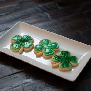 Puede incluir: Cuatro galletas de azúcar en forma de trébol verde en un plato rectangular blanco. Las galletas están decoradas con glaseado verde y chispas verdes.