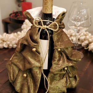 Op de afbeelding: Een fles wijn is versierd met een decoratieve olijfgroene fluwelen flessenhoes. De hoes heeft gouden accenten, waaronder een decoratieve strik en kleine gouden kralen. De gouden dop van de fles heeft een reliëf bloemmotief.