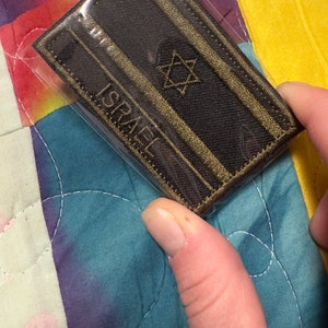 May include: A brown and gold embroidered patch with the word "ISRAEL" and a six-pointed star in the centre.