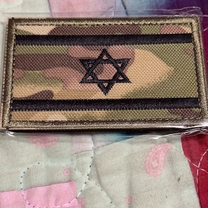 May include: A camouflage fabric patch with a black Star of David in the center. The patch has a brown border.