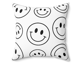 May include: White square pillow with a repeating pattern of black smiley faces. The faces have black eyes and curved smiles. The pillow is a decorative item, suitable for a bed or couch.