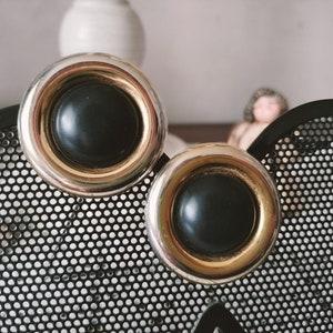 May include: A pair of large, round clip-on earrings. Each earring features a black center, a gold-colored inner ring, and a silver-colored outer ring. The earrings are displayed on a black, perforated metal surface.