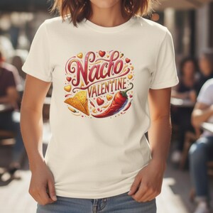 May include: Cream-colored t-shirt featuring a colorful graphic that reads "Nacho Valentine." The design includes a cartoon taco, a red chili pepper, and heart shapes. The shirt is made of a soft material.