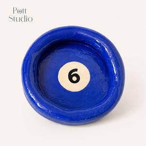 Op de afbeelding: Een helderblauwe, cirkelvormige schaal met een glanzende afwerking. De schaal heeft een witte cirkel met het nummer 6 in zwart, die doet denken aan een biljartbal. Het is een uniek decoratief item.