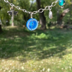 May include: A silver chain necklace with various charms. The charms include a dragonfly, a blue and white circular stone, a spiral, and a teal glass bead. The necklace is displayed against a blurred green background.
