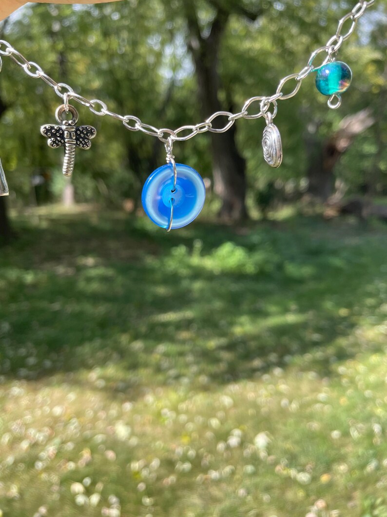 May include: A silver chain necklace with various charms. The charms include a dragonfly, a blue and white circular stone, a spiral, and a teal glass bead. The necklace is displayed against a blurred green background.