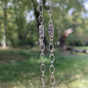 May include: A silver necklace with two strands of linked chain. Each strand features a decorative cylindrical bead with a spiral design. The background is a blurred view of green trees and foliage.
