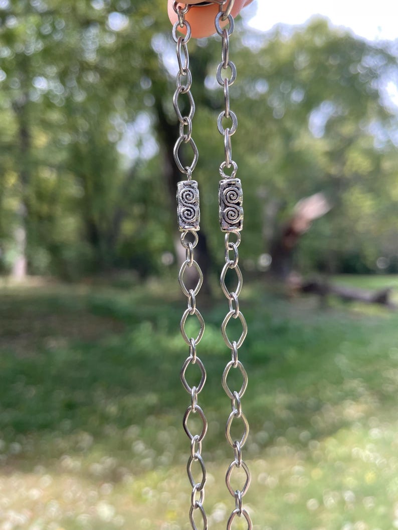 May include: A silver necklace with two strands of linked chain. Each strand features a decorative cylindrical bead with a spiral design. The background is a blurred view of green trees and foliage.