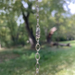May include: A silver chain with a decorative cylindrical bead featuring a spiral design. The chain is composed of small, oval and circular links. The background is a blurred view of green foliage and trees.