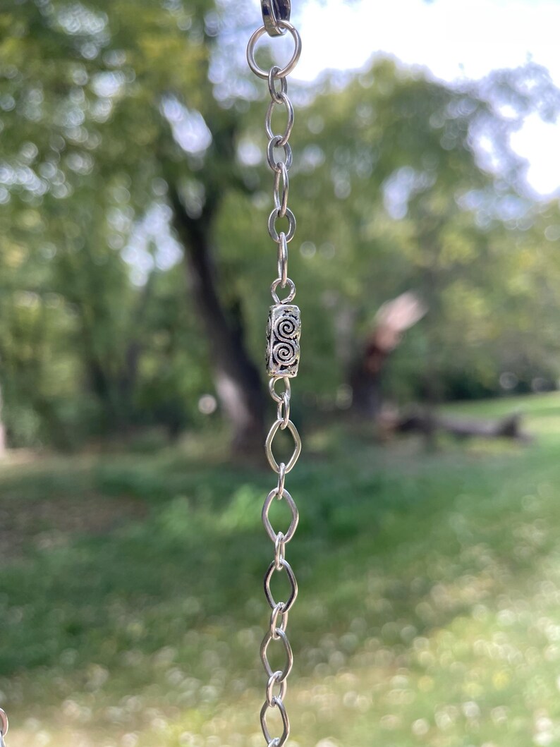 May include: A silver chain with a decorative cylindrical bead featuring a spiral design. The chain is composed of small, oval and circular links. The background is a blurred view of green foliage and trees.