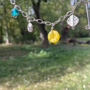 May include: A silver chain necklace with various charms, including a key, a blue bead, a yellow bead, and a silver spiral. The necklace is displayed against a blurred green background of trees and grass.