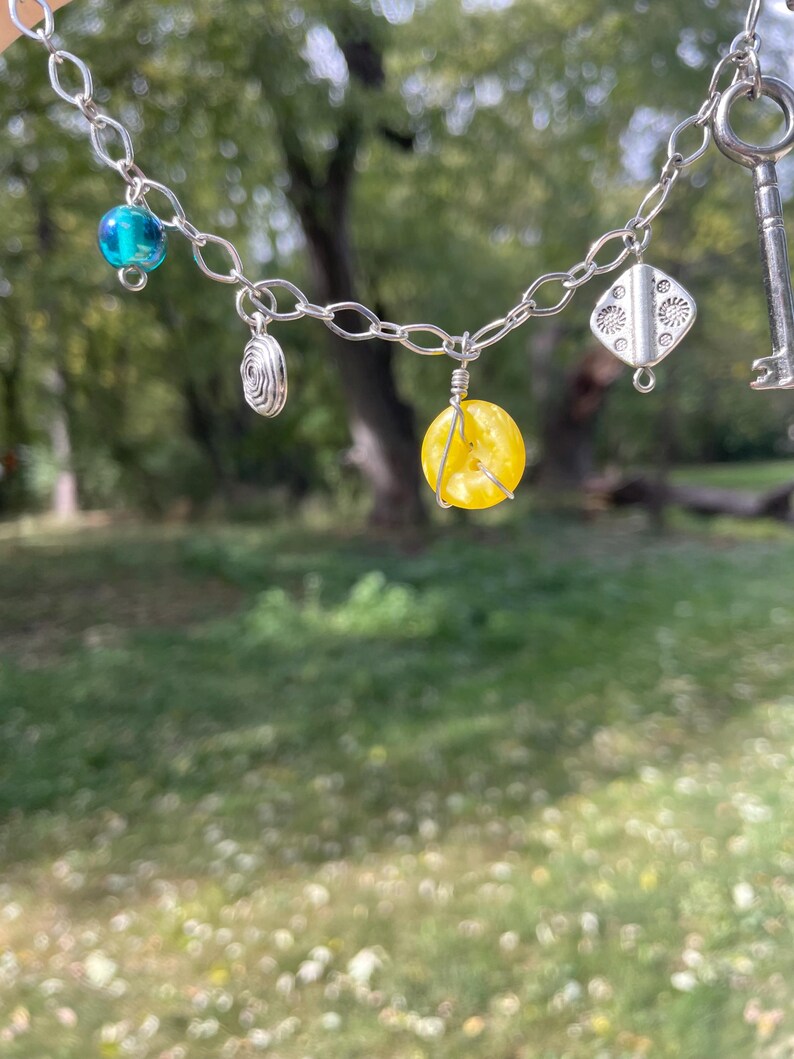 May include: A silver chain necklace with various charms, including a key, a blue bead, a yellow bead, and a silver spiral. The necklace is displayed against a blurred green background of trees and grass.