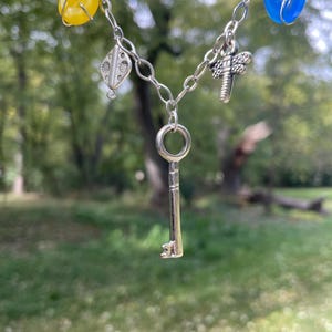May include: A silver-toned necklace with a key pendant and various charms. The necklace features yellow and blue beads, a dragonfly charm, and a decorative silver charm. The key pendant hangs in the center, creating a unique and eye-catching design.