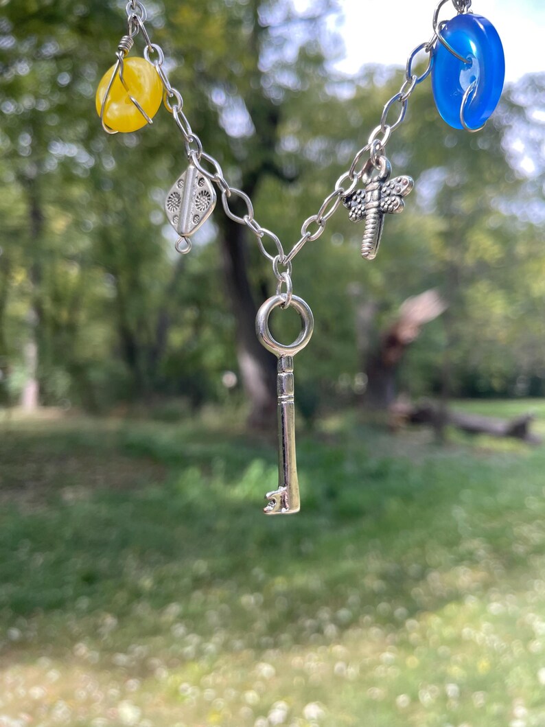 May include: A silver-toned necklace with a key pendant and various charms. The necklace features yellow and blue beads, a dragonfly charm, and a decorative silver charm. The key pendant hangs in the center, creating a unique and eye-catching design.