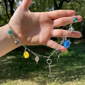 May include: Silver chain necklace with various charms, including a key, spiral, leaf, and colorful beads in blue, yellow, and green. The necklace is displayed against a blurred green background.