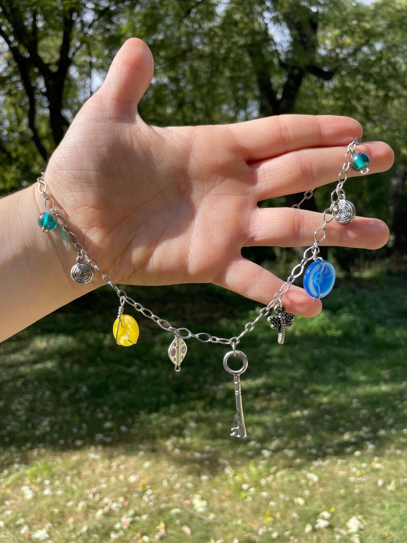 May include: Silver chain necklace with various charms, including a key, spiral, leaf, and colorful beads in blue, yellow, and green. The necklace is displayed against a blurred green background.