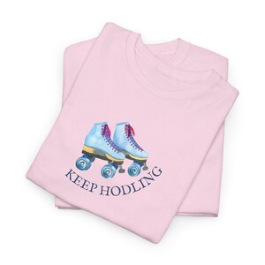 Puede incluir: Camiseta rosa con un par de patines de ruedas azules y el texto "KEEP HODLING" en azul marino.