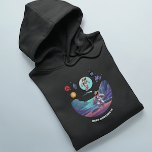 May include: Black hoodie with a graphic of two astronauts in space with cryptocurrency symbols. The text "KEEP HODLING IT" is printed on the hoodie.