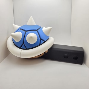 May include: A blue and white spiky shell with a black rectangular base. The shell has a blue geometric pattern with white accents and spikes. The shell is resting on a black rectangular object with two circular holes.
