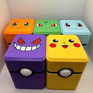 May include: Six colorful, square storage boxes with character faces painted on the lids. The boxes are orange, green, blue, purple, and yellow. Each box has a white circular button on the front.