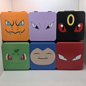 May include: Six square wooden boxes, each painted with a different cartoon character face. The boxes are orange, purple, black, green, blue, and red. The faces have various expressions and colors, with white and black accents.