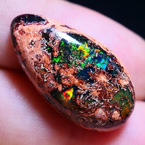 Może przedstawiać: Kropelkowaty opal szlachetny o iryzującym połysku w kolorze brązowym, czarnym i zielonym. Opal jest osadzony w surowej, naturalnej macierzy.