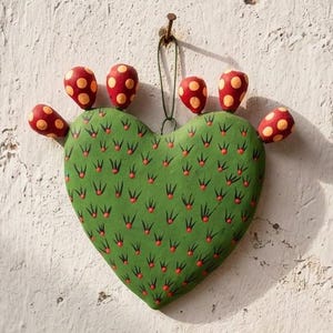 May include: A heart-shaped cactus wall hanging painted green with red spines and six red, yellow-spotted fruit accents attached to the top edge. The object is suspended by a thin string from a nail on a textured, off-white vertical surface.