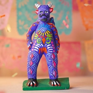Könnte beinhalten: Eine violette Tonfigur eines gehörnten Wesens, das auf einem grünen rechteckigen Sockel steht. Die Figur hat einen aufgemalten Schnurrbart, spitze Ohren und komplexe, bunte Muster in Gelb, Weiß, Rosa und Blau auf ihrem gesamten Körper.