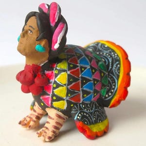 Puede incluir: Una figura de cerámica pintada a mano que muestra una cabeza humana con cabello oscuro y pendientes turquesa sobre un cuerpo de pavo. El pavo tiene una base negra con triángulos geométricos coloridos y una cola de color naranja y rojo.