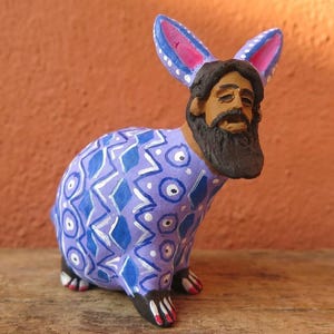 Puede incluir: Una pequeña figura de conejo con rostro humano que presenta barba y bigote de color marrón oscuro. El cuerpo del conejo está pintado en tonos violeta y azul con un patrón repetitivo de círculos y diamantes blancos. Tiene orejas largas con centros rosas.