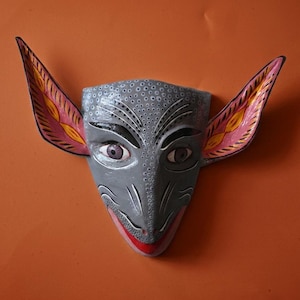 May include: A hand-painted wall mask featuring a grey, textured face with large, pointed ears. The ears have pink and yellow accents, while the eyes are purple. The mask has a pointed nose, black eyebrow details, and a red mouth at the bottom edge.