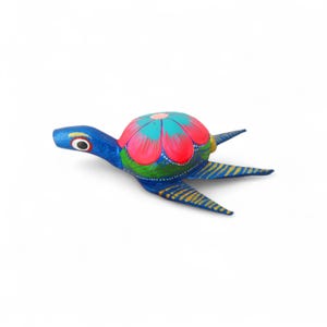 Może przedstawiać: Ręcznie malowana drewniana figurka żółwia. Żółw ma niebieski korpus, głowę i płetwy. Skorupa jest ozdobiona różowymi, turkusowymi i zielonymi wzorami kwiatowymi. Płetwy mają żółte paski. Oko żółwia jest czarne z białym odblaskiem.