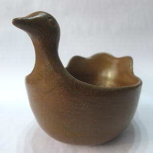 May include: A brown ceramic duck-shaped bowl. The bowl has a rounded base and a scalloped rim. The duck's head and neck form a handle, with a detailed beak and eye. The bowl is likely handmade and has a rustic, textured finish.
