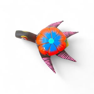 Może przedstawiać: Ręcznie malowana drewniana figurka żółwia. Skorupa żółwia ma kwiatowy wzór w kolorach pomarańczowym, niebieskim i białym. Płetwy są różowe z czarnymi paskami, a głowa jest ciemnobrązowa z żółtymi i czerwonymi akcentami.