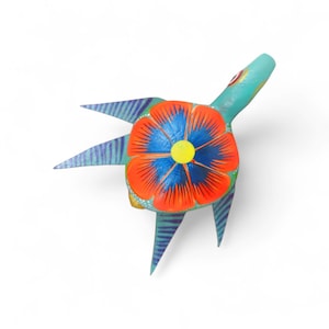 Może przedstawiać: Ręcznie malowana drewniana figurka żółwia z dużym kwiatowym wzorem na grzbiecie. Kwiat ma czerwone płatki, niebieski środek i żółty środek. Żółw jest pomalowany w odcieniach niebieskiego i zielonego, z paskowanymi płetwami.