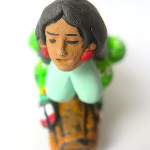May include: A hand-painted clay figurine featuring a person with black hair, red earrings, and a light green top. The figure wears a green patterned garment and is positioned atop a textured brown base with dark markings, captured from a high angle.