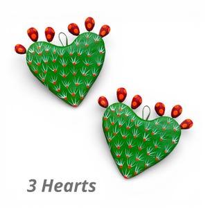 Könnte beinhalten: Zwei herzförmige Kaktus-Ornamente. Die grünen Kaktusherzen haben weiße Stacheln und rote Punkte. Jeder Kaktus hat rot-orangefarbene fruchtartige Akzente. Der Text "3 Hearts" befindet sich unten.