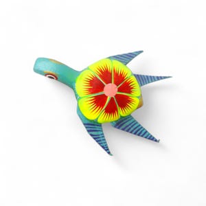 Może przedstawiać: Ręcznie malowana drewniana figurka żółwia. Żółw pomalowany jest w odcieniach turkusu, żółci, czerwieni i niebieskiego. Skorupa ozdobiona jest dużym kwiatowym wzorem. Płetwy są niebieskie z paskami.